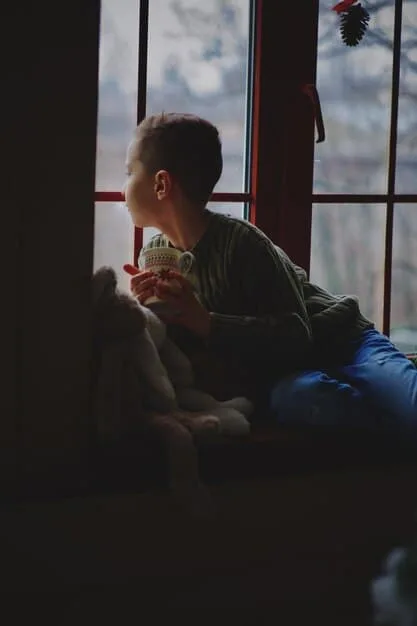 A person sitting in a comfortable chair, holding a cup of tea, and looking out of a window at a rainy cityscape. The scene conveys a sense of quiet contemplation and introspection.