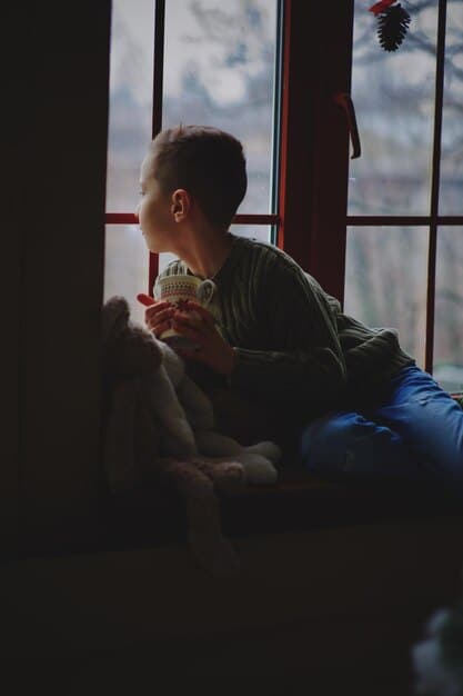 A person sitting in a comfortable chair, holding a cup of tea, and looking out of a window at a rainy cityscape. The scene conveys a sense of quiet contemplation and introspection.