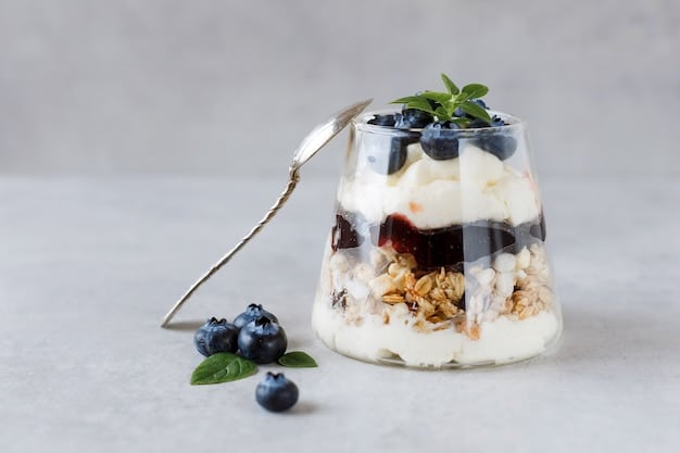 A layered Greek yogurt parfait in a clear glass. The layers consist of Greek yogurt, fresh blueberries, granola, and a drizzle of honey. A spoon is placed beside the glass, inviting consumption.